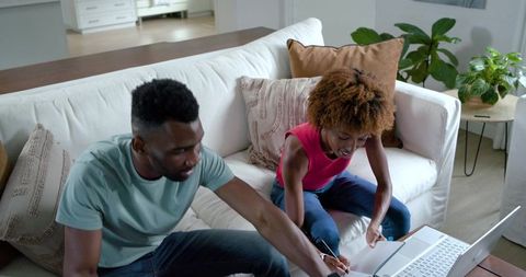 Black couple collaborating on laptop while taking notes on cozy living room sofa