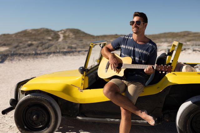 Smiling caucasian man in sunglasses sitting on beach buggy playing guitar on sunny beach. beach stop off on summer holiday road trip.