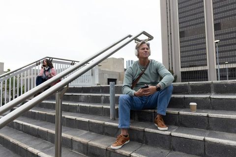 Middle-aged man listening to music on headphones while using smartphone on urban steps