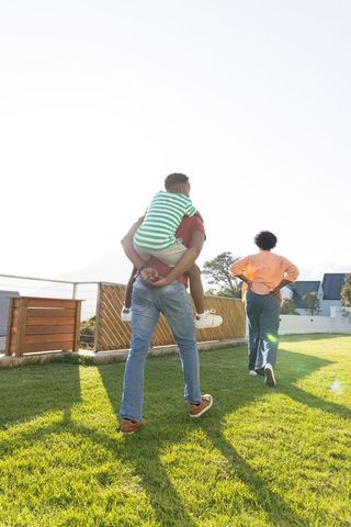 African american family walking on sunlit lawn carrying child piggyback in backyard
