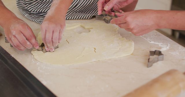 Hands are cutting shapes in rolled-out cookie dough using cookie stencils on a floured table. Use in articles or blogs about homemade baking, cooking activities, family fun, and teamwork in the kitchen.