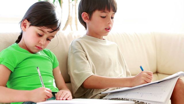 Young girl and boy sitting on couch, concentrating on their homework. Scene suggests at-home learning or after-school study routine. Useful for educational materials, family lifestyle concepts, and advertisements for home furniture or educational supplies.