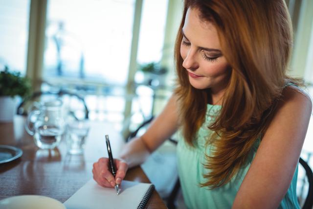 Woman sitting at table and writing on notepad in cafÃ©