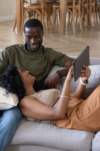 Reclining diverse couple sharing tablet on sofa, relaxing together in cozy living room