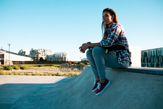 Caucasian woman sitting on a wall at a skatepark, enjoying the summer sun. She is dressed casually in a plaid shirt, jeans, and sneakers, holding a skateboard. The urban backdrop and clear sky suggest a relaxed, leisurely day. Ideal for use in lifestyle, recreation, and urban culture themes.