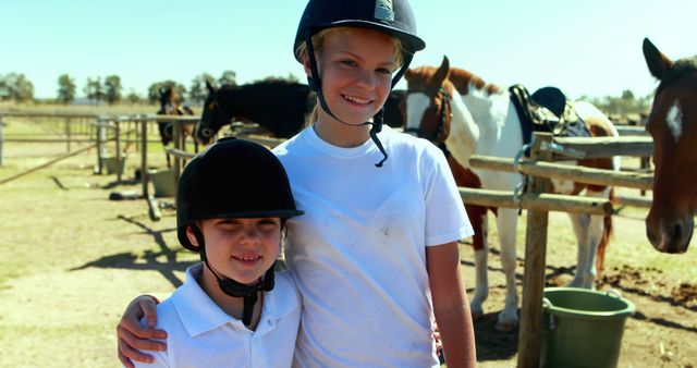 Two children wearing helmets and white shirts are enjoying horseback riding at a farm. They are smiling and appear to be siblings, standing in front of several horses on a sunny day. Ideal for use in content related to family activities, outdoor sports, equestrian hobbies, summer camps, or rural living.