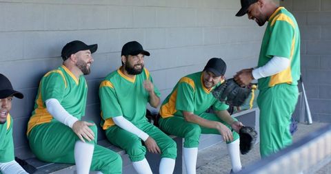 Male baseball team laughing and chatting in dugout wearing green gold uniforms with gloves