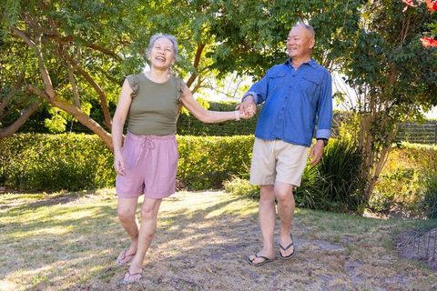 Senior asian couple holding hands laughing and walking in sunlit backyard wearing flip-flops
