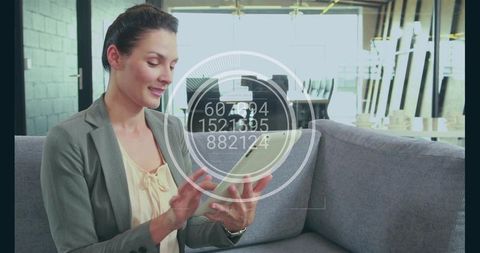 Businesswoman tapping tablet on sofa in modern coworking lounge with futuristic hud