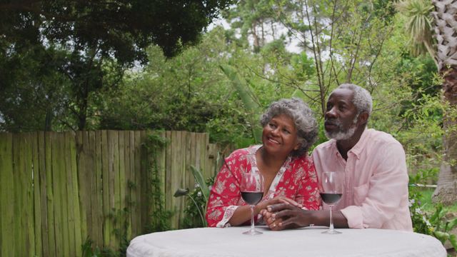 A senior African American couple is smiling and looking away while sitting at a table with glasses of red wine in their garden. They are enjoying quality time together during quarantine due to the coronavirus pandemic. This video can be used for topics related to family, elderly well-being, romance, and lockdown experiences. Perfect for articles, blogs, and advertisements focused on love, care, and health measures during challenging times.