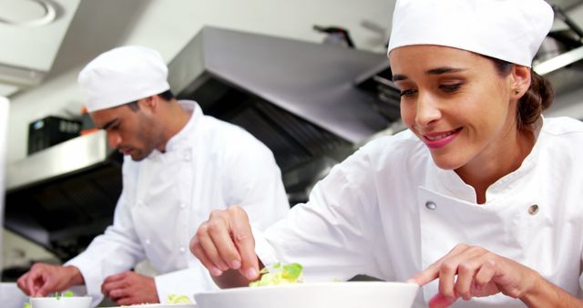 Two chefs in a commercial kitchen are preparing dishes with concentration. They are both wearing white uniforms and chef hats. The focus is on the chef smiling while delicately preparing a dish. Great for stories about culinary arts, restaurant industry, cooking classes, and teamwork in professional settings.