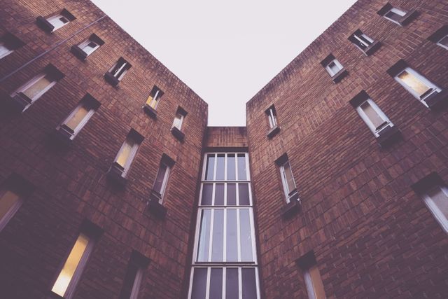This image captures a low angle view of a modern brick building against the sky. The symmetrical design of the windows complements the geometric structure, creating an abstract and visually appealing perspective. Ideal for use in architecture blogs, urban design websites, real estate promotions, and modern-day architectural publications.
