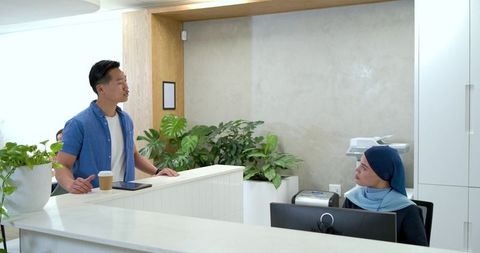 Asian man leaning on reception desk, receptionist in headscarf assisting in neutral lobby