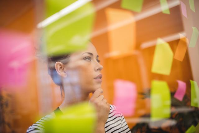 Woman in professional environment analyzing colorful sticky notes on glass wall. Ideal for portraying concepts of brainstorming, planning sessions, strategy meetings, or business ideas development. Useful for business presentations, creative industry content, and teamwork illustrations.