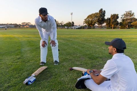Diverse cricket teammates resting and chatting on outfield during sunset practice