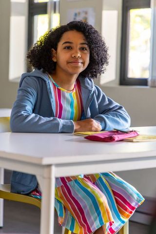 Preteen girl sitting at classroom desk in striped rainbow dress studying attentively