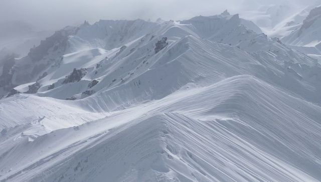 Wind-Sculpted Alpine Ridge Showing Snow Cornices and Striations across Misty Glacier Peaks