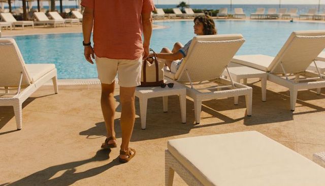 Poolside couple retreat man walking past tote and sunglasses on sunlit oceanfront deck