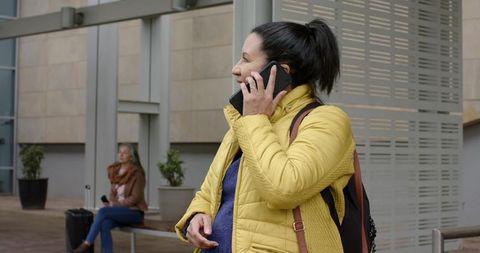 Senior Woman Calling on Phone at Transit Hub Wearing Yellow Jacket and Backpack