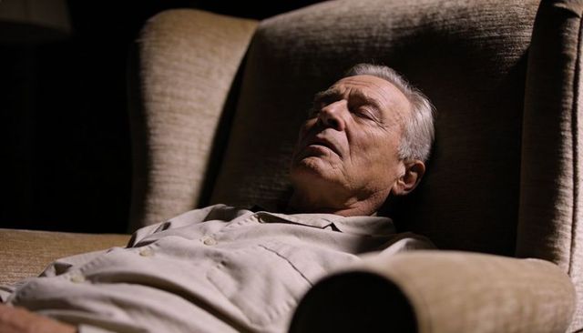 Reclining senior man dozing in highback beige armchair warm dim lighting intimate mood