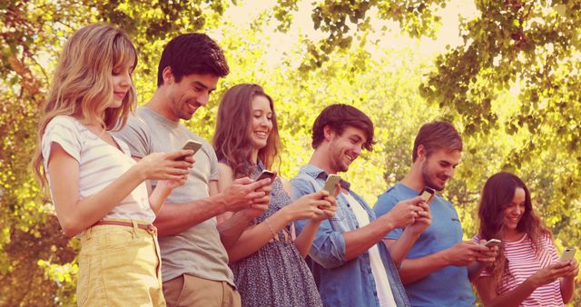 Group of young people standing outdoors in a row, interacting with their smartphones under a sunny sky, surrounded by trees. Ideal for topics related to social media, technology, youth culture, connectivity, and communication in modern society.