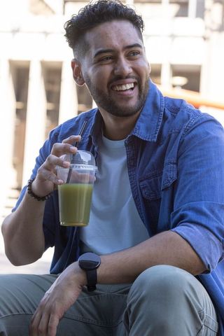 African american man smiling and holding green smoothie while sitting on urban plaza steps