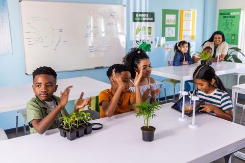 Diverse students clapping while using tablet and exploring mini wind turbines and seedlings