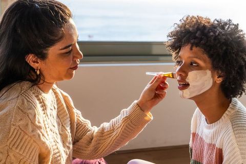 Diverse adult applying face mask to teen by sunlit window for cozy pampering moment
