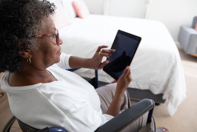 This image depicts an elderly African American woman sitting in a wheelchair and using a digital tablet in her bedroom. It can be used to illustrate themes of independence, accessibility, and the use of modern technology by older adults. Suitable for articles or advertisements related to senior living, healthcare, technology for the elderly, and inclusive design.