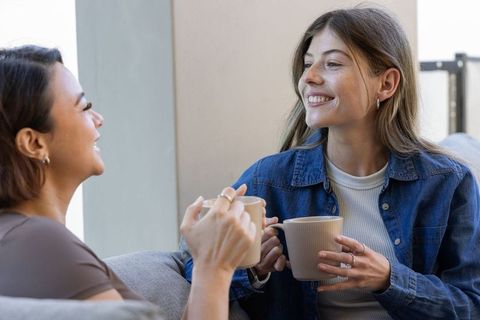 Smiling diverse female friends chatting over coffee on sunlit sofa, casual conversation
