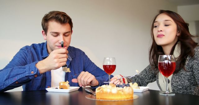 Young couple sitting at table, eating cake and enjoying their time together. Glasses of red drink add a festive touch. Ideal for concepts such as home dining, relationships, happiness, enjoying life's sweet moments.