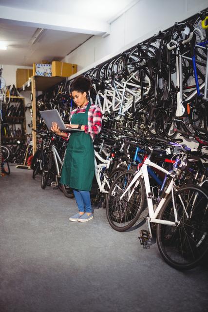 Female mechanic using laptop in bicycle workshop. She is standing surrounded by various bicycles, and appears to be assessing data or ordering parts. Useful for themes related to technology in trades, bicycle maintenance, and female professionals in technical fields.