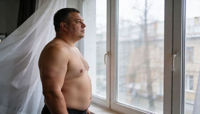 Shirtless middle-aged man standing and gazing out apartment window, contemplative mood