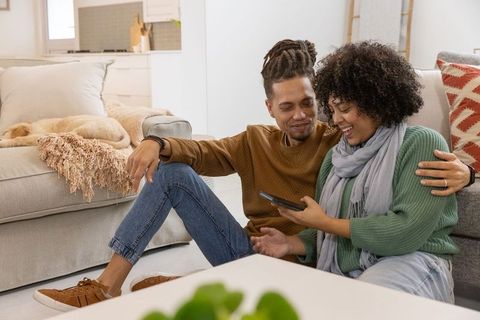 Couple laughing and relaxing sharing smartphone on living room floor with sleeping dog