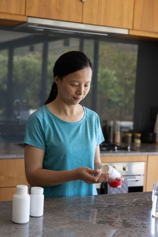 Mature asian woman pouring pills at kitchen counter with water for supplement routine