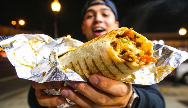 Holding and offering grilled burrito in saucy foil on urban night street with bokeh lights