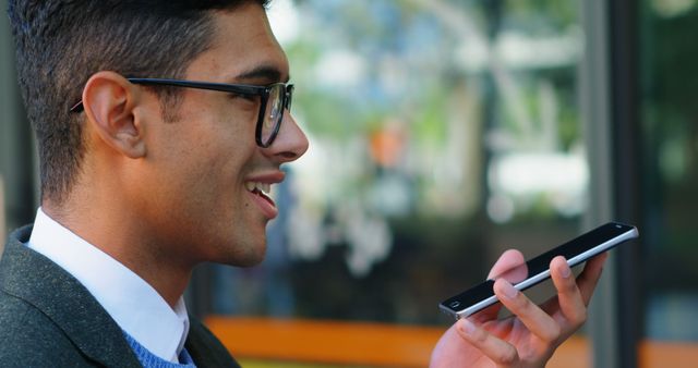 Young Asian man using a voice assistant on smartphone while standing outside. Man is wearing glasses and appears relaxed. Suitable for concepts relating to modern communication, technology, smart devices, or casual outdoor activities.