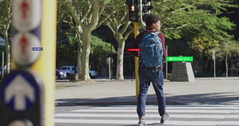 Man in Maroon Sweater with Backpack Crossing Urban Street at Traffic Light