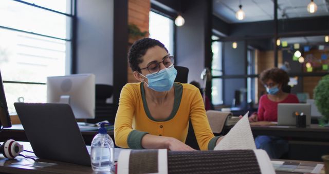 Female designer wearing a protective mask sitting at workstation in a modern coworking space. She's focused on reviewing fabric samples, with a laptop and sanitizer bottle nearby. Perfect for depicting safe workplace practices during a pandemic, modern office environment, and creative professions.