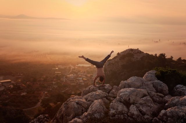 This image captures a man performing a handstand on a rocky cliff as the sun rises over a fog-filled valley. Ideal for themes of adventure, fitness, and motivation, or background for travel and outdoor activity campaigns.
