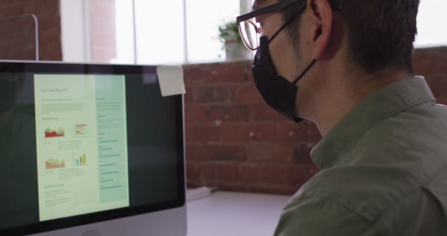 Man in casual attire wearing glasses and face mask, analyzing financial data displayed on computer screen in a modern brick-walled office. Useful for themes related to business, finance, remote work, productivity, workplace safety, and technology.