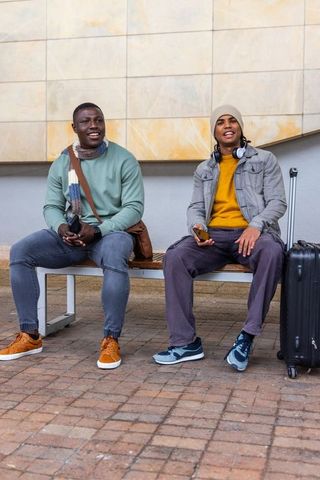 Two men sitting and chatting at transit bench with suitcase, headphones and winter layers