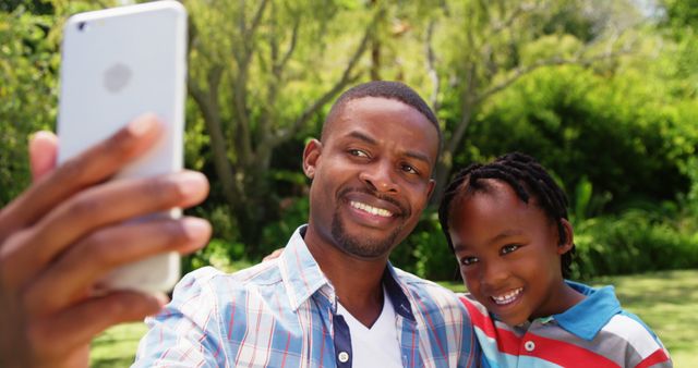 Father and son are smiling and taking a selfie together in a lush, green garden. Perfect for themes of family bonding, outdoor activities, happiness, and father-son relationships. Suitable for use in family event promotions, Father's Day activities, social media content, and lifestyle blogs.