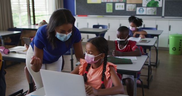 Diverse female teacher helping schoolgirl using laptop, all wearing face masks. children in primary school during coronavirus covid 19 pandemic.