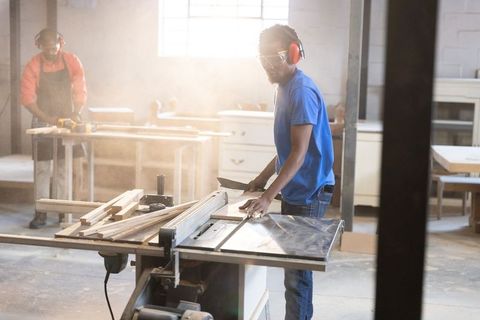 African american carpenter guiding plank through table saw in dusty woodworking workshop