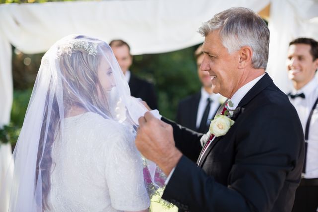 Father lifting veil from bride's face during wedding ceremony, capturing a heartfelt moment. Ideal for use in wedding planning materials, family celebration promotions, or articles about wedding traditions and special moments.
