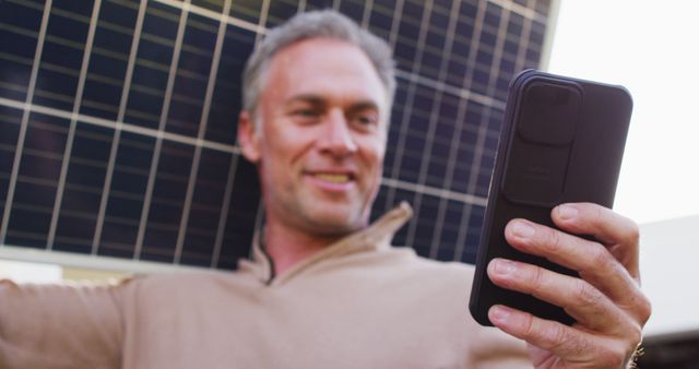 Happy caucasian man holding solar panel, using smartphone and smiling. Spending time at home alone.