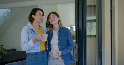 Female friends sharing coffee and laughing by sliding door in modern home interior