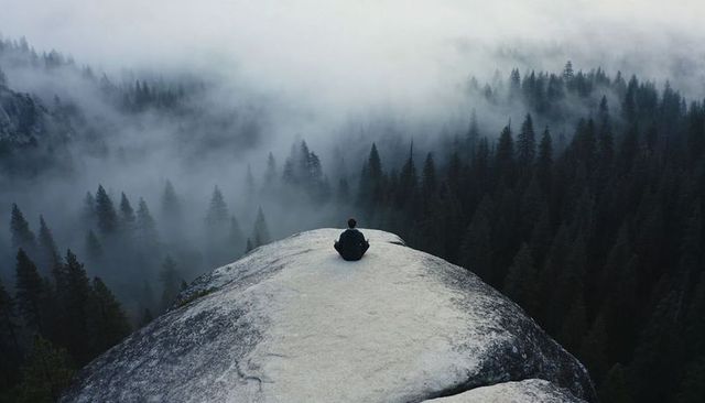 Solitary hiker sitting on granite outcrop overlooking misty conifer forest valley at dawn