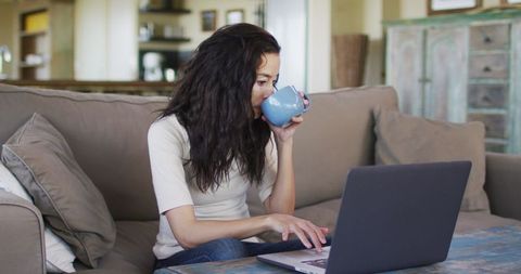 Woman on sofa enjoying coffee while working on laptop